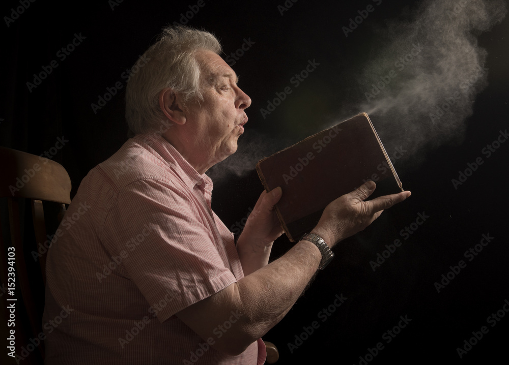 Mature man blowing dust off an old book, taken on a black background